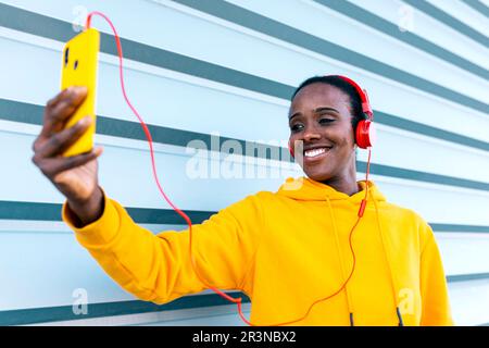 Positive young female in headphones smiling and making video call on smartphone while standing on street against blue lined wall Stock Photo