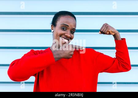 Portrait of content African American female model looking at camera and smiling while pointing at bicep and standing against blue wall in city Stock Photo
