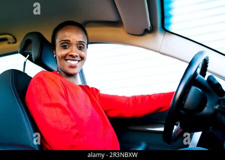From below positive adult African American woman with dark hair in red sweater smiling and looking at camera while sitting in modern car on sunny day Stock Photo