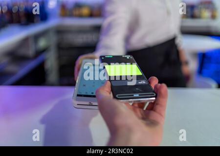 Unrecognizable male bartender showing bill on POS machine while anonymous customer paying with mobile phone at counter against blurred bar interior Stock Photo