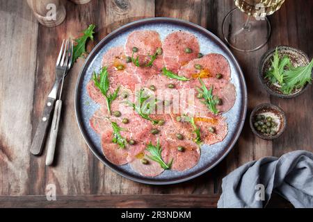 Marbled beef carpaccio with arugula and capers on gray plate, with white wine, wooden background, top view Stock Photo