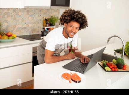 Happy Black Guy Cooking Near Laptop Posing In Kitchen Indoor Stock Photo