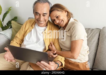 Retirement lifestyle and technologies concept. Happy senior spouses video calling using laptop, sitting on couch at home Stock Photo