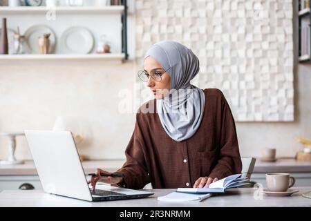 Young muslim woman in hijab freelancer working from home in kitchen. He sits focused on the laptop and makes notes in the notebook. Stock Photo
