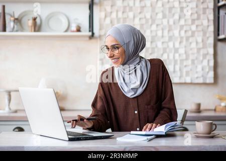 Arab young female student studying remotely from home. He sits at the kitchen table at the laptop and writes the summary in a notebook. Stock Photo