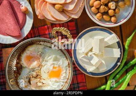 Traditional homemade Turkish breakfast with fried eggs in a copper pan, vegetables and meat, top view on the dining table in the morning sun, hearty b Stock Photo