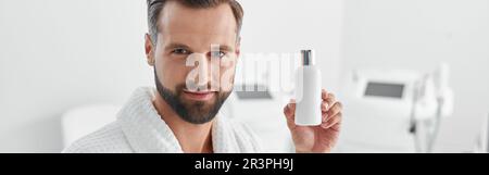 Attractive mature man holds bottle of cosmetic product standing in cosmetologycal clinic Stock Photo
