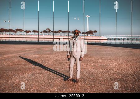 A stylish black man in a light-colored suit and striped tie stands on a brown cobblestone floor, with flag poles in the background and a glimpse of th Stock Photo