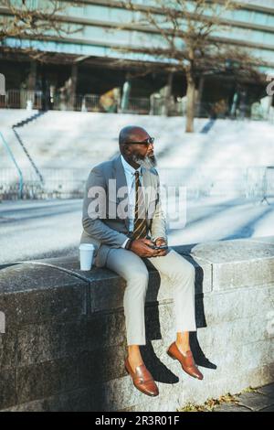 A bald man with a full white-bearded face holding a smartphone, has his cup of coffee next to him, dressed in a cream suit with a yellow striped tie, Stock Photo