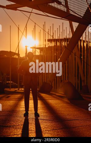 Vertical; In Lisbon, a black man stands before a white tensile structure, bathed in golden hour light. Dramatic backlighting creates captivating long Stock Photo