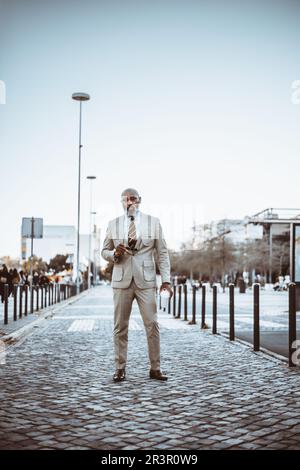 Vertical capture of a stylish bearded black man with a shaven head, dressed in a beige full suit, holding a cup of coffee in the middle of a touristic Stock Photo