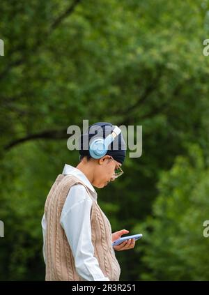 Young girl using phone to listen music on her headphones in the park. Technology wireless headphones for listening the music via smart mobile phone. Y Stock Photo