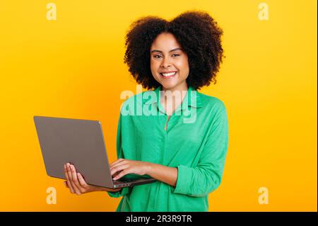 Positive beautiful brazilian or african american curly haired woman, dressed in a green shirt, holding an open laptop in hand, looking at camera, smiling, standing over isolated orange background Stock Photo