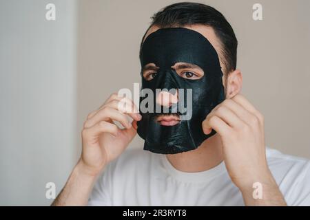 Handsome man applying black face mask on his face, receiving spa treatments. Grooming himself. Facial treatment, cosmetology. Facial skin care Stock Photo