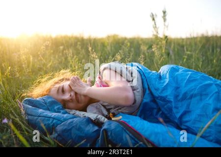 The girl is dissatisfied with scratching mosquito bites, child sleeps in a sleeping bag on the grass in a camping trip. Eco-frie Stock Photo