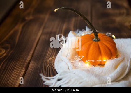 Orange pumpkin wrapped in a warm white plaid with lights of garlands on a wooden table with copyspace. Autumn mood, halloween, h Stock Photo