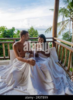 Couple waking up in bed looking out over ocean during sunrise at wooden hut moutains of Thailand Stock Photo
