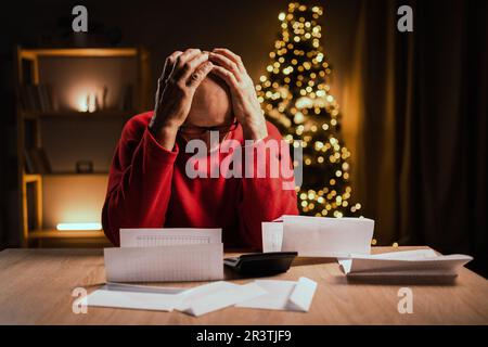 Worried mature men cheeking mail or bills late night in home office during christmas holidays, holding head with hands, finance problem and headache. Stock Photo