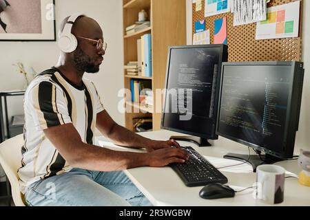 African American programmer in wireless headphones developing computer codes on computer sitting at his workplace in office Stock Photo