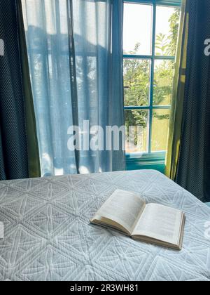 Open book lies on a white quilted spread on the bed. Sunlight shines and green garden is visible through the bedroom windows. Stock Photo
