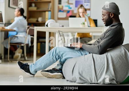 African American man sitting on comfortable bag chair and working online on laptop in office Stock Photo