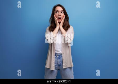 surprised puzzled 30s dark haired woman in shirt and jeans on blue background Stock Photo