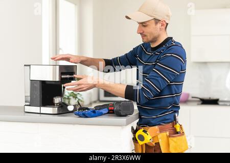 Professional young repairman repairs a coffee maker. Handsome worker in uniform repairing coffee machine in a workshop. Stock Photo