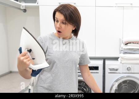 Woman looking with surprise at bottom of broken iron in laundry room with washing machine on background Stock Photo