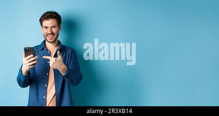 Handsome smiling man pointing finger at smartphone and looking pleased at camera, recommending app, standing on blue background Stock Photo