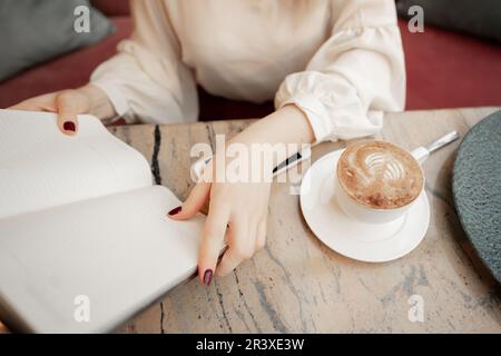 Cropped image of girl making notes in open notepad while sitting at table and resting in modern cozy cafe Stock Photo