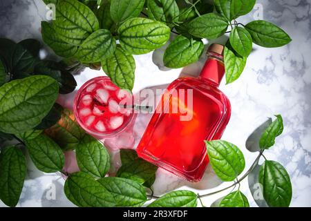 A bottle of red alcohol, next to it in an old-fashioned glass is a cocktail with liquor and ice.  On a marble background, surrounded by green leaves o Stock Photo
