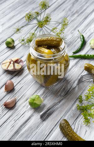 Pickled cucumbers in an open jar, gherkin on a fork and garlic on a light wooden background. Homemade canned cucumbers. Stock Photo