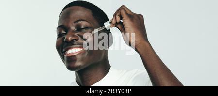 Young black man smiling while applying beauty oil to his melanated face in a studio. Happy youthful man proudly practicing his skincare routine with a Stock Photo