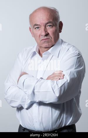 Confident senior man in white shirt crossing hands on chest and looking at camera while standing against gray background. Self c Stock Photo
