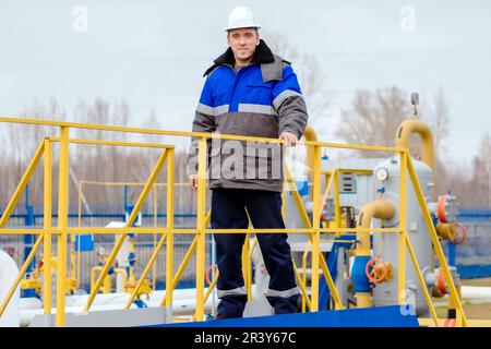 Portrait of worker in white helmet and winter pea jacket at industrial gas facility. Middle-aged Caucasian man stands in full growth on territory of gas distribution station. Genuine worker. Stock Photo
