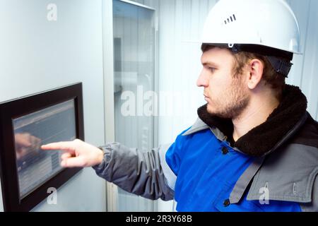 Engineer in hard hat and winter pea coat presses his finger on touch screen of modern production equipment indoors. Setting up automatic control system. Gas distribution station operator monitors gas flow readings. Photo. side view. Stock Photo