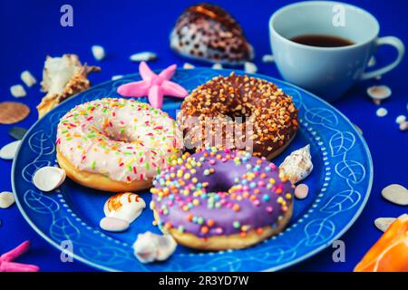 Assorted donuts in a plate on a blue background. Around them are symbols of the sea - shells, pebbles and stones. The concept of pleasure and relaxati Stock Photo
