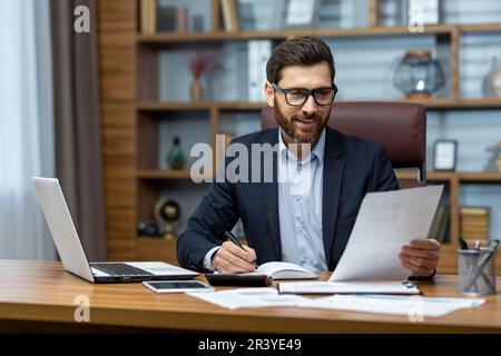 Mature successful financier working inside office at workplace with contracts and papers businessman behind paper work reading financial report, experienced analyst satisfied with achievement results Stock Photo