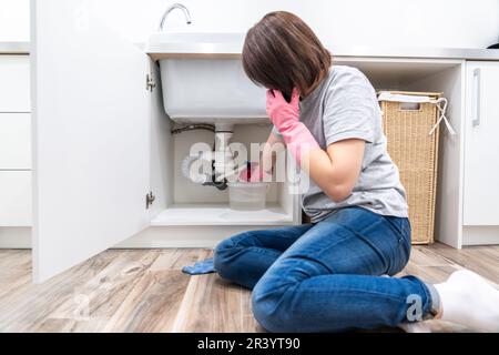 Woman sitting near leaking sink in laundry room calling for help Stock Photo