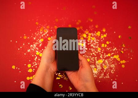 Beautiful female hands hold a phone over a red background with silver sparkles and gold round confetti. View from above. Blank s Stock Photo