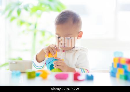 Adorable Asian baby boy playing with colorful blocks toy in white sunny bedroom. Cute baby with educational toys. Child building tower. Stock Photo