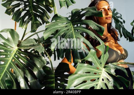 Beautiful brunette female expecting a child stands in monstera leaves Stock Photo
