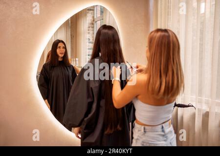 Blonde hairdresser cuts her female client hair in a beauty salon. Pretty brunette young woman looks into a huge mirror when her Stock Photo