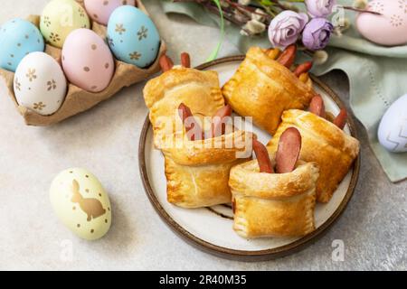 Buns in the form of an Easter rabbit from sausage and cheese in a yeast dough with colored eggs on a gray stone background. Trad Stock Photo