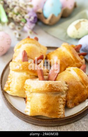 Buns in the form of an Easter rabbit from sausage and cheese in a yeast dough with colored eggs on a gray stone background. Trad Stock Photo