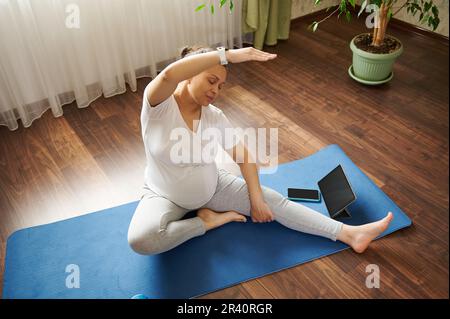 Top view of a pregnant woman doing yoga on a blue exercise mat in front of digital gadget, repeating after coach online Stock Photo
