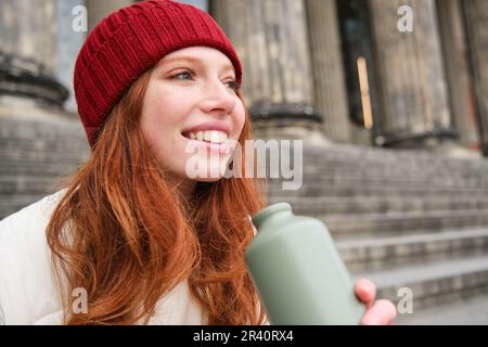 Smiling redhead girl in red hat, sits on stairs near building with flask, drinks water and looks happy Stock Photo