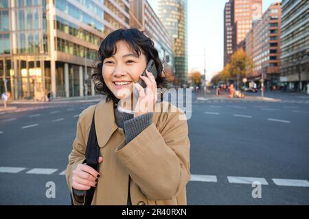 Smiling asian girl makes a phone call, stands on an empty street, calling someone on telephone, waiting for friend in city, goin Stock Photo