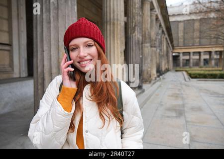 Smiling redhead female tourist talks on mobile phone and walks around city. Happy student in red hat calls friend, stands on str Stock Photo