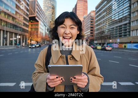 Enthusiastic asian girl with digital tablet, smiling and looking amazed, happy with something good, holding digital tablet, stan Stock Photo